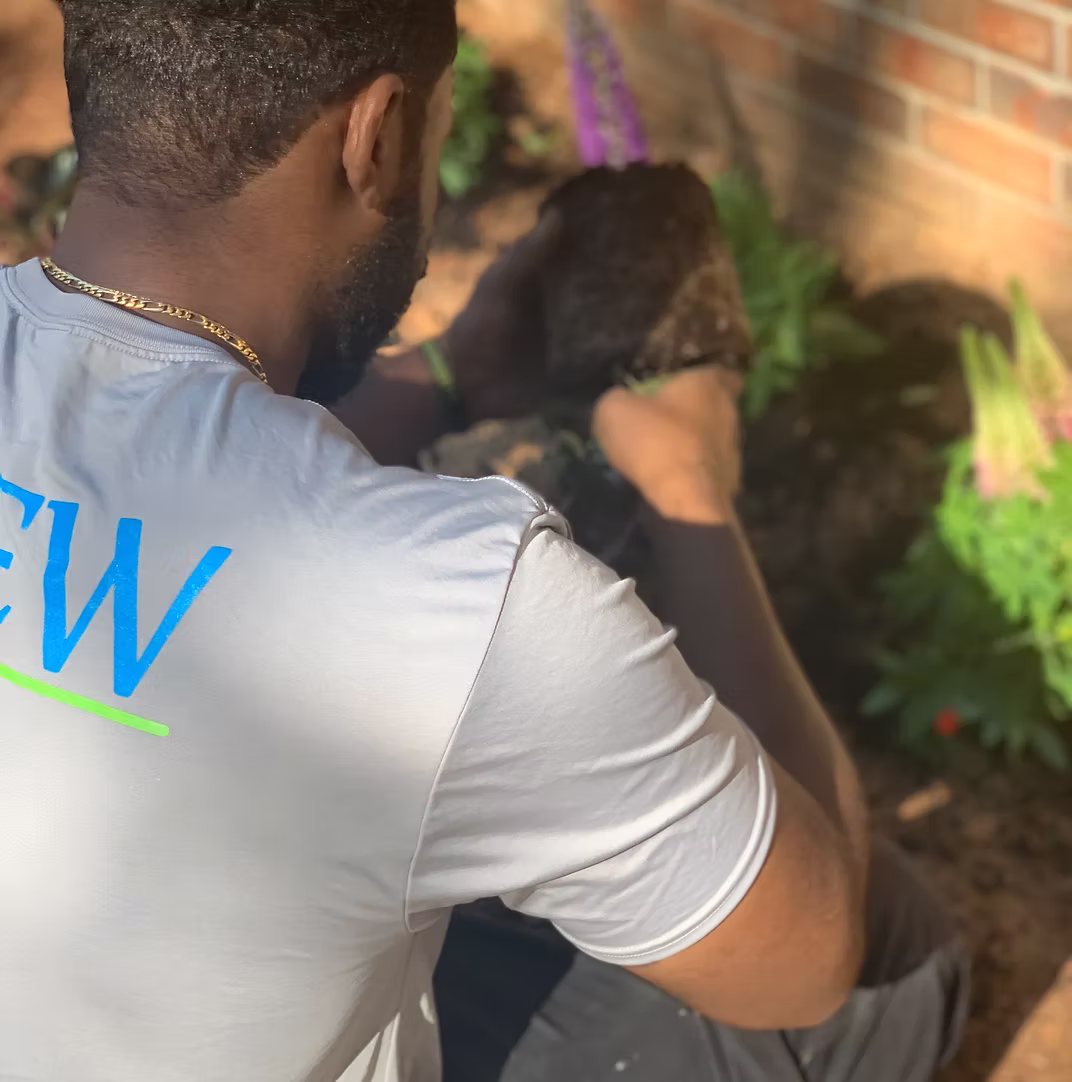 Person in white t-shirt with "NEW" text sitting near brick wall and plants